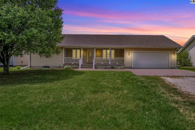 Light yellow ranch home with a covered porch, white railings, and a two-car garage under a colorful sunset.