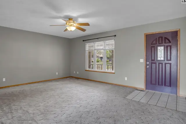Spacious living room with gray walls, textured carpet, ceiling fan, and a purple front door.