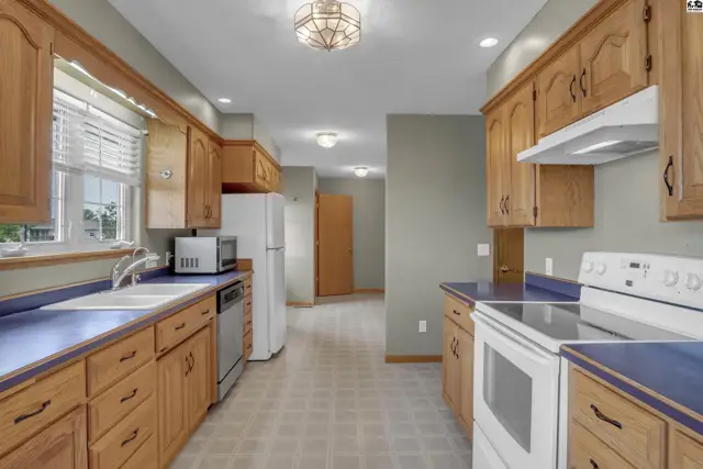 Spacious kitchen with natural wood cabinets, blue countertops, white appliances, and tiled flooring.