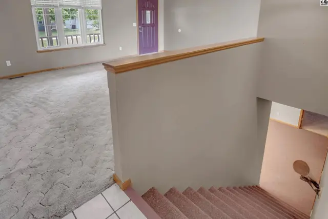 Spacious living area with textured carpet, large windows, and a purple front door, overlooking a carpeted staircase.