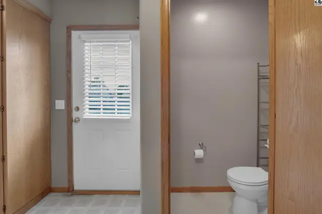 Hallway with a door to the outside and a bathroom featuring a toilet and shelving.