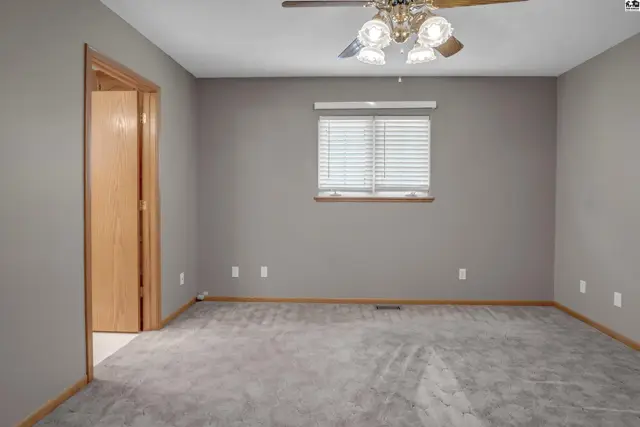 Spacious gray bedroom with plush carpet, a ceiling fan, and a single window with blinds.