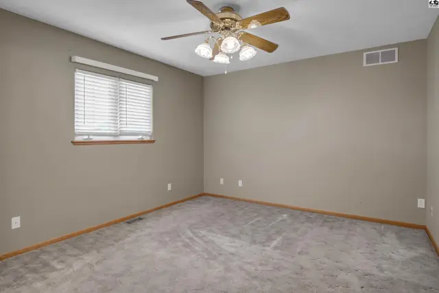 Spacious bedroom with neutral carpet, beige walls, and a wood-bladed ceiling fan.
