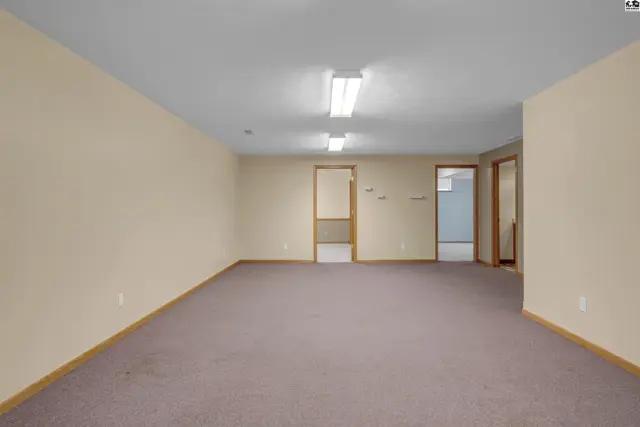 Spacious room with beige walls, brown carpet, and fluorescent lighting, featuring multiple doorways.