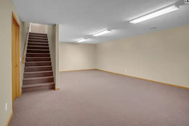 Spacious, carpeted basement with neutral walls, a wooden staircase, and bright overhead lighting.