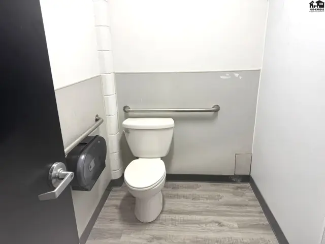 Accessible bathroom with toilet, grab bars, paper towel dispenser, and wood-look flooring.