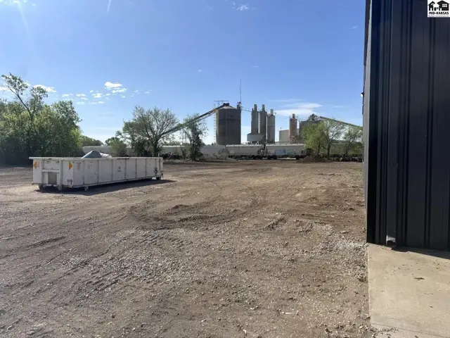 Industrial site with a large dumpster, gravel lot, and distant concrete silos under a clear sky.