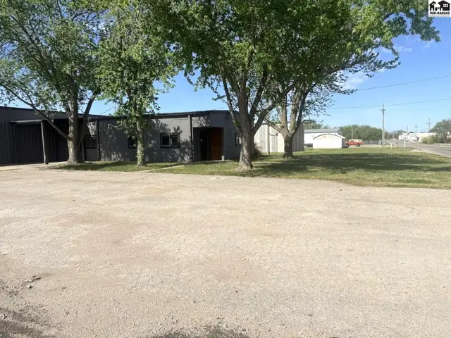 Gray metal commercial building with large trees and a gravel parking lot.
