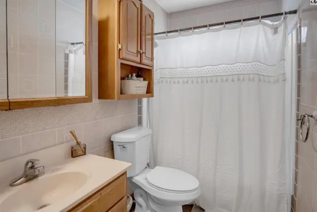 Bright bathroom with a vanity, toilet, and white shower curtain against tiled walls.