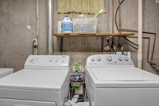 Laundry room with two white top-load washers and dryers, concrete walls, and a wooden shelf.