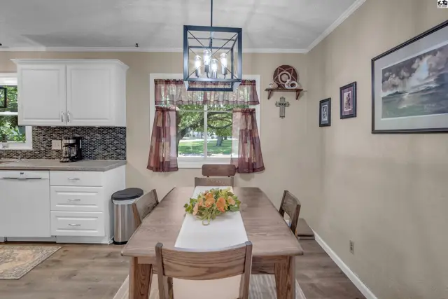 Dining area with a wooden table, chairs, and a modern chandelier, adjacent to a kitchen with white cabinets.