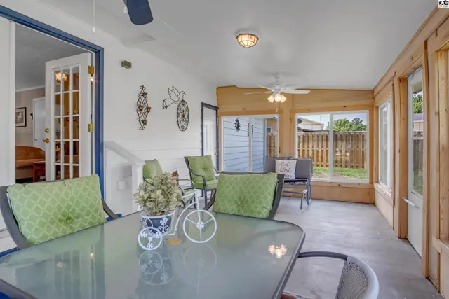 Sunroom with glass-top table, green-cushioned chairs, and wood-paneled walls overlooking a fenced yard.