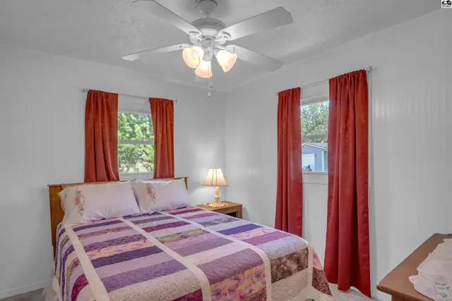 Cozy bedroom with a queen bed, colorful quilt, white ceiling fan, and red curtains.