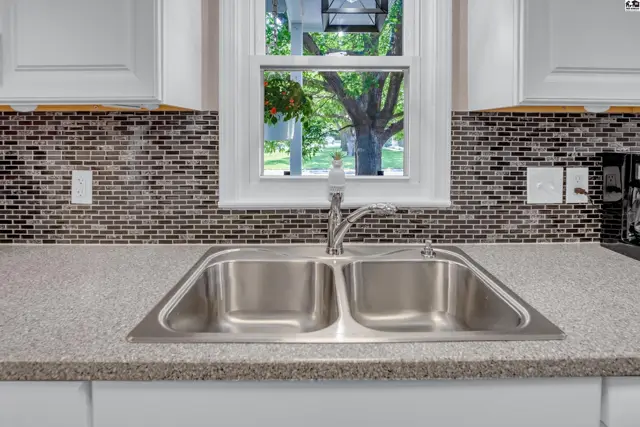 Kitchen sink with granite counters and a mosaic tile backsplash overlooks a green, tree-filled yard.