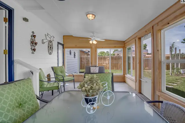Sunroom with glass-top table, green upholstered chairs, wood paneling, and large windows overlooking a backyard.