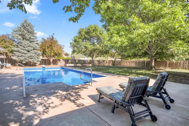 Rectangular swimming pool with lounge chairs on a concrete patio surrounded by a wooden fence and mature trees.