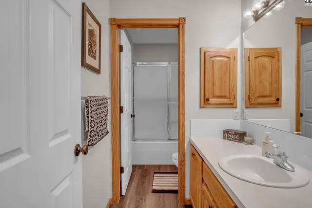 Bright bathroom with a white vanity, wood cabinets, and a glass-enclosed shower visible through the doorway.
