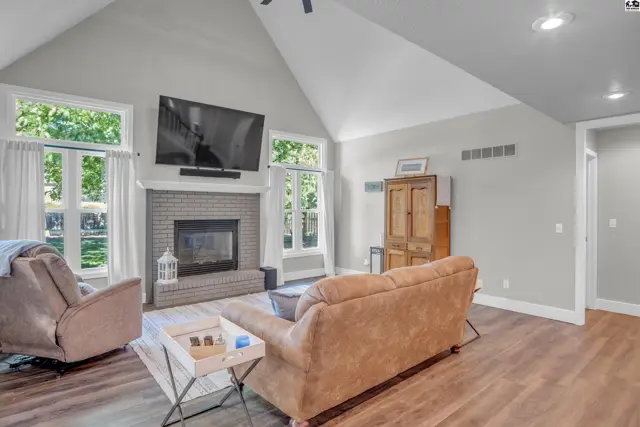 Spacious living room with vaulted ceiling, brick fireplace, large windows, and wood-look flooring.