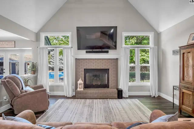 Spacious living room with vaulted ceiling, brick fireplace, large windows, and hardwood floors.
