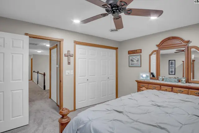 Spacious bedroom with a ceiling fan, wood dresser, mirrored vanity, and carpeted floor.