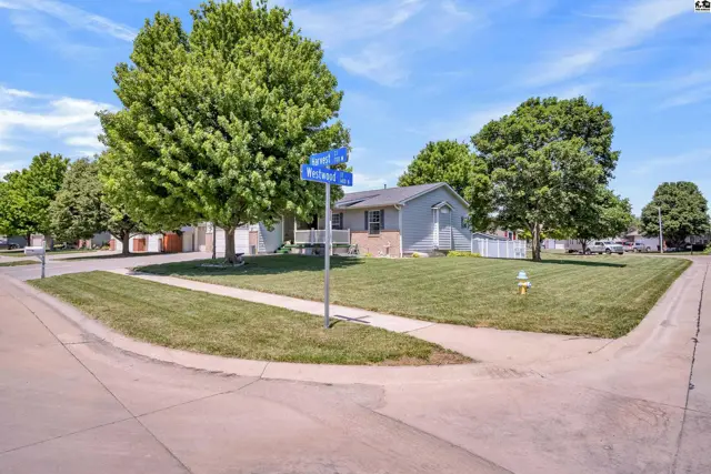 Corner lot ranch home with light gray siding, brick accents, and mature trees on a sunny day.