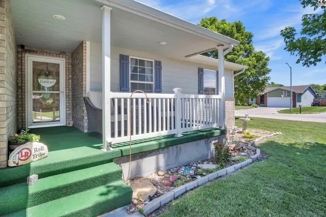 Covered front porch with white railing, blue shutters, and green carpeted steps leading to a brick entryway.