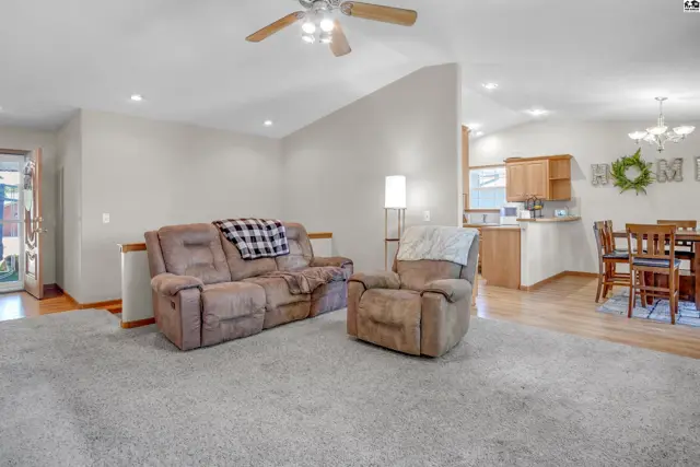 Spacious living room with vaulted ceiling, plush carpet, and adjacent dining area with wood cabinets.