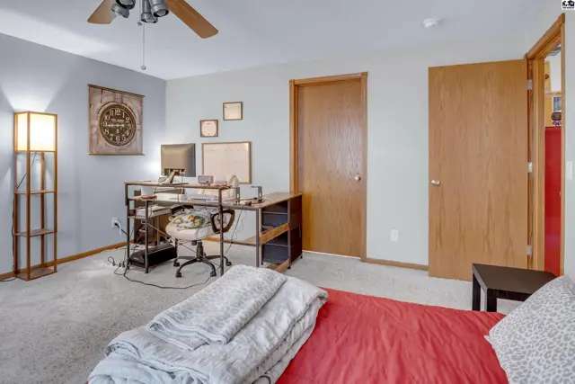 Spacious bedroom with a desk, dual doors, and a floor lamp, featuring light gray walls and carpet.
