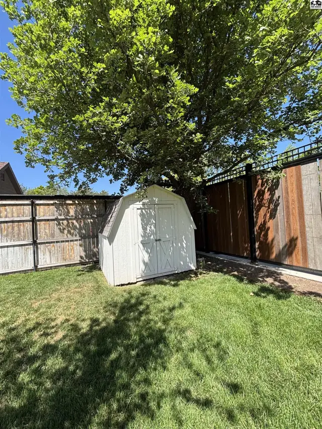 White barn-style shed sits on a green lawn beneath a large tree, bordered by wooden fences.
