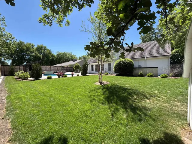 Spacious backyard with a swimming pool, lush green lawn, mature trees, and a white stucco home.