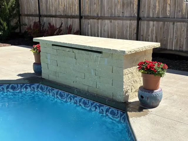 Stone waterfall feature with planters and a pool in a backyard setting.