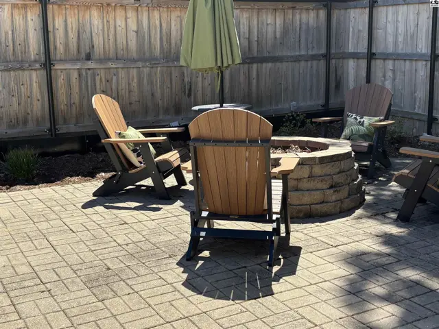 Paved patio with Adirondack chairs surrounding a stone fire pit under a wooden fence.