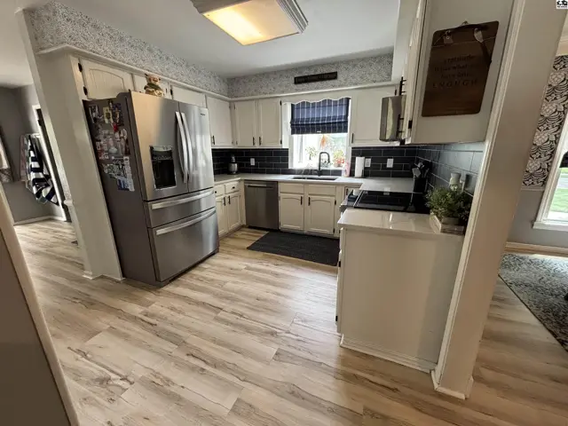 Bright kitchen with white cabinets, stainless steel appliances, dark tile backsplash, and light wood-look flooring.