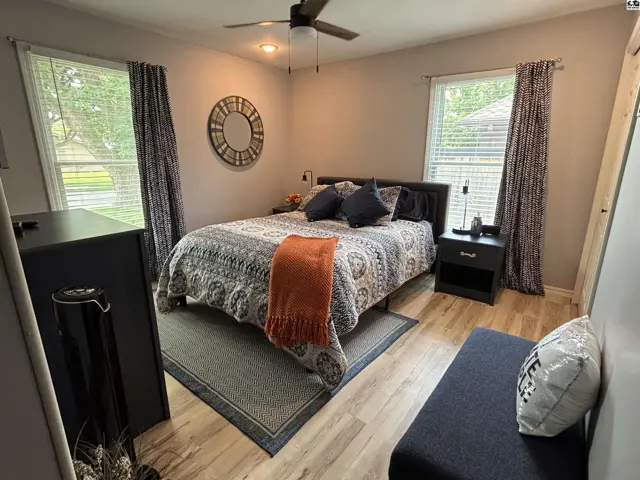 Cozy bedroom with a queen bed, patterned quilt, wood-look flooring, and large windows.
