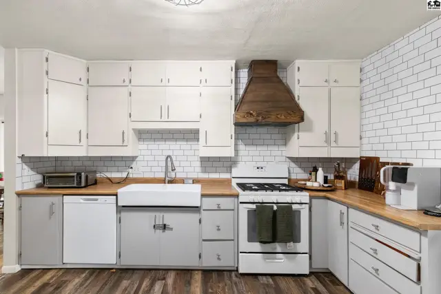 Bright kitchen with white subway tile backsplash, light gray cabinets, butcher block counters, and a rustic wood range hood.