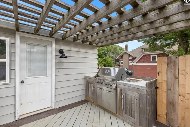 Outdoor kitchen with stainless steel grill and rustic wood cabinetry under a wooden pergola.