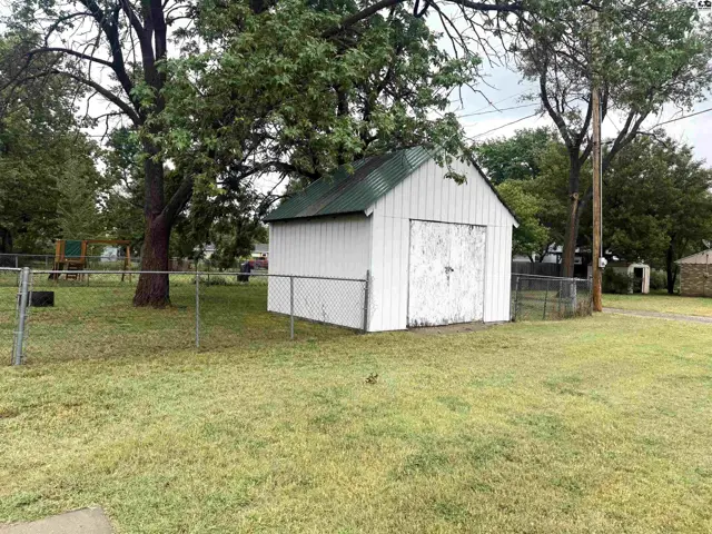 White shed with green metal roof and double doors sits in a grassy yard with a chain-link fence.