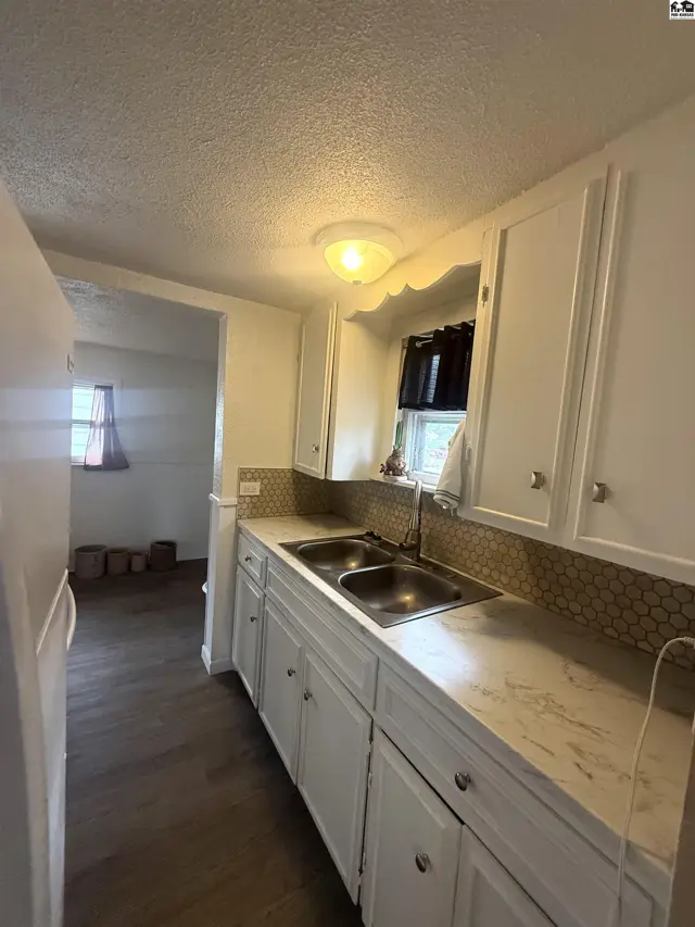 Bright kitchen with white cabinets, stainless steel sink, and mosaic tile backsplash, opening to a living area.