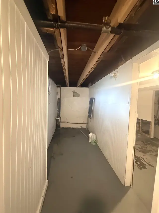 Unfinished basement hallway with white paneled walls, exposed wooden beams, and gray concrete floor.