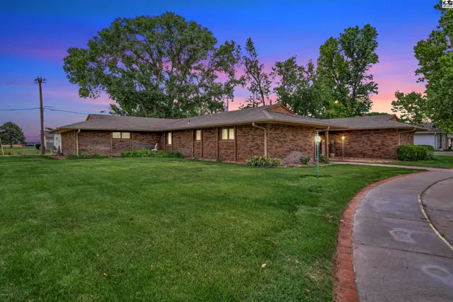 Sprawling brick ranch home with lush green lawn and mature trees under a colorful twilight sky.