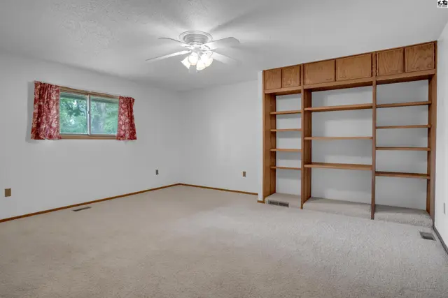 Spacious room with plush carpet, a large built-in wooden bookshelf, and a ceiling fan.