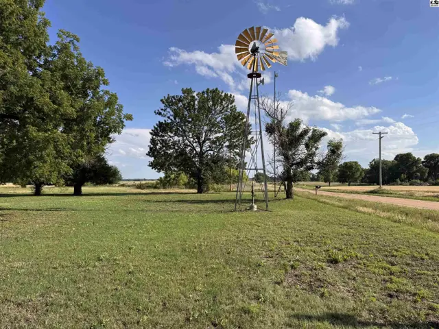 Rustic metal windmill stands in a grassy field with mature trees under a bright blue sky.