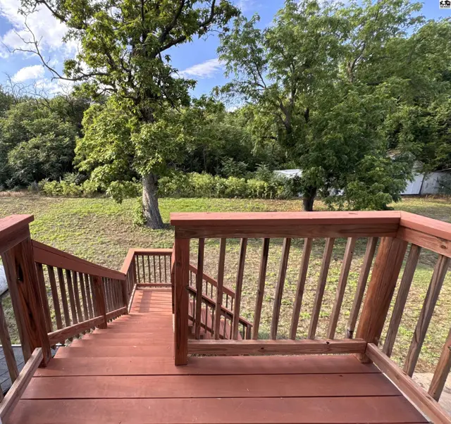Wooden deck stairs with red stain lead down to a grassy yard with mature trees.