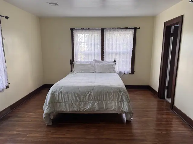 Spacious bedroom with a queen bed, hardwood floors, and large windows with white lace curtains.
