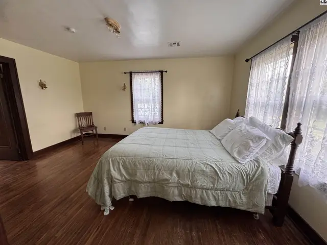 Spacious bedroom with a queen bed, hardwood floors, and lace-curtained windows.