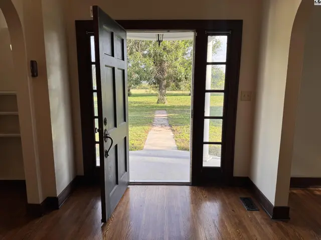 Spacious entryway with dark wood floors, a grand open door, and large windows revealing a tree-lined path.