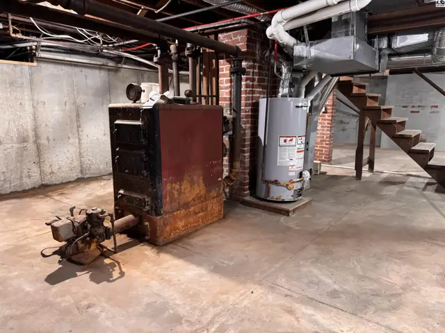 Basement utility area with a large, rusty boiler, water heater, and unfinished concrete floor.
