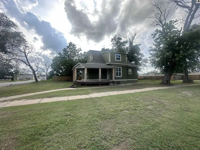 Green house with a wide front porch, white trim, and mature trees on a grassy lot.