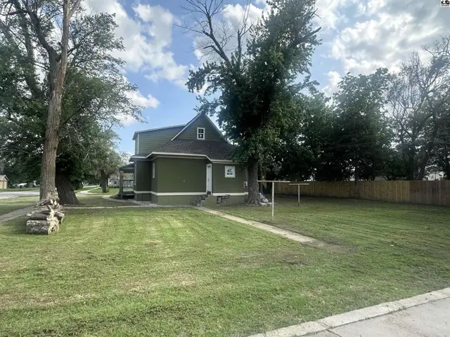 Green siding house with a large yard and mature trees under a cloudy sky.