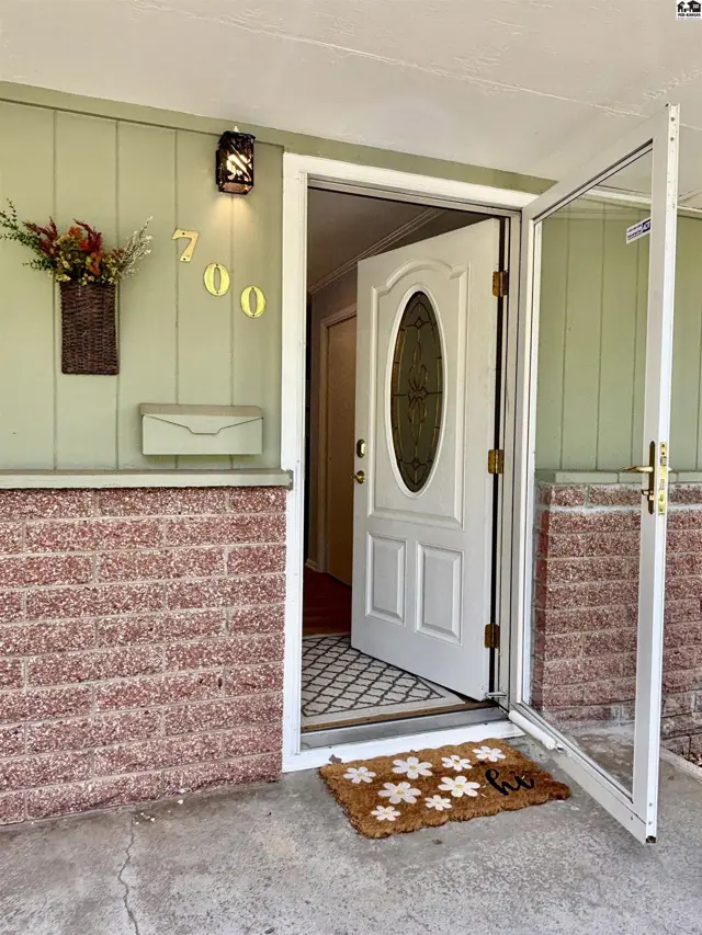 Light green siding and red brick exterior with an open white door, oval glass, and floral doormat.